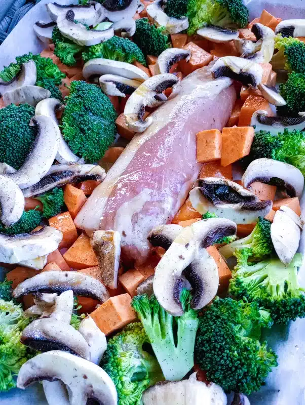 pork and mushroom recipe. pork loin surrounded by broccoli, sweet potatoes, and mushrooms on a baking sheet before going into the oven